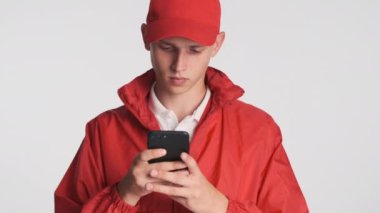 Kırmızı şapkalı ve ceketli genç teslimatçı beyaz arka planda akıllı telefon kullanıyor.