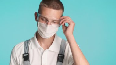 Üniformalı genç müteahhitin portresi tıbbi maske ve koruyucu gözlükleri çıkarıyor mavi arka planda kameraya güvenle bakıyor.