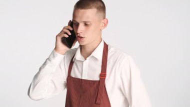 Üniformalı genç çekici garson beyaz arka planda telefonla konuşuyor.