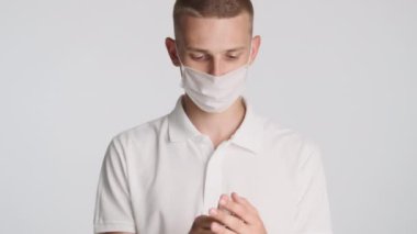 Tıbbi maskeli genç adam beyaz arka planda antiseptikle ellerini yıkıyor. Güvenlik kavramı
