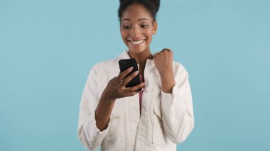Çekici, neşeli, Afro-Amerikalı kız akıllı telefon kullanıyor ve renkli arka planda kameraya seviniyor.