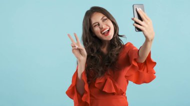 Kırmızı elbiseli çekici esmer kız mutlu bir şekilde barış jesti yapıyor. Renkli arka planda akıllı telefondan selfie çekiyor.