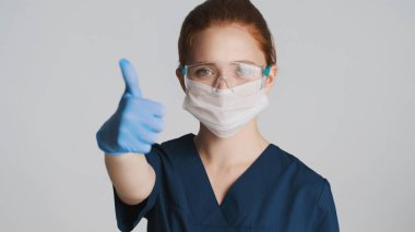 Koruyucu gözlüklü, tıbbi maskeli ve eldivenli genç bayan doktor kamerada baş parmağını gösteriyor. Aferin ifade.