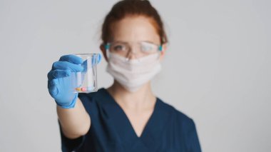 Koruyucu gözlüklü, maskeli ve eldivenli genç profesyonel doktor kamerada güvenli bir şekilde tıbbi matarayı gösteriyor.