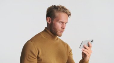 Çekici, heyecanlı, sarışın, sakallı adam beyaz arka planda mutlu bir şekilde akıllı telefondan video izliyor.