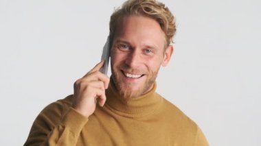 Çekici, gülümseyen, sarışın, sakallı adam beyaz arka planda akıllı telefondan mutlu bir şekilde konuşuyor. Kendine güvenen adam.