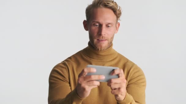 迷人而兴奋的金发大胡子男人 一边玩着智能手机 一边对着白底笑 — 图库视频影像