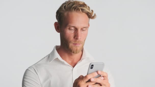 ハンサムなブロンドひげを生やしたビジネスマンでシャツの中で熱心に仕事をスマートフォンと笑顔上の白い背景 — ストック動画