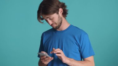 Mavi tişörtlü çekici sakallı adam şüpheli bir şekilde renkli arka planda akıllı telefon kullanıyor.