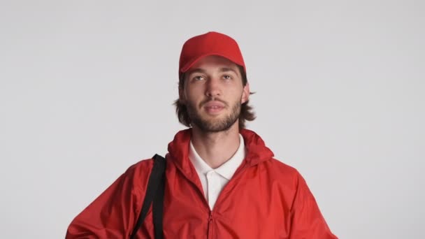 有魅力的送货员头戴红帽子 身穿夹克 背着背包 高高兴兴地举起食指 用大拇指在白色背景上摆出姿势 — 图库视频影像