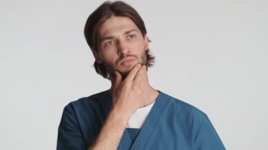 Genç ve ciddi bir erkek doktor beyaz arka plan üzerinden kameraya kızgın bir şekilde bakıyor. Düşünceli ifade