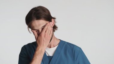 Genç sakallı erkek doktor, beyaz arka planda kameraya poz veriyor. Yorgun ifade