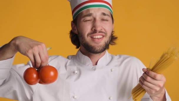 迷人而快乐的意大利厨师 身穿制服 手持西红柿和通心粉 在色彩艳丽的背景下 在镜头前欢快地歌唱 — 图库视频影像