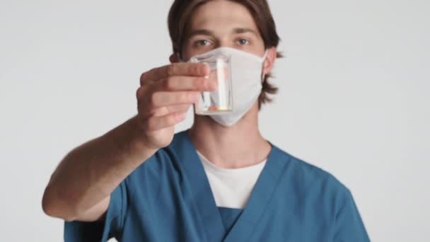 白い背景にカメラに自信を持って医療薬を示すマスクの若い魅力的な男性医師 安全コンセプト — ストック動画