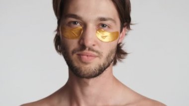 Gözlerinin altında altın bantları olan çekici sakallı adam beyaz arka planda kameraya güvenle poz veriyor.