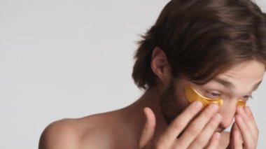 Gözlerinin altında altın bantlar olan yakışıklı sakallı adam beyaz arka planda kameraya baş parmak hareketi yapıyor.