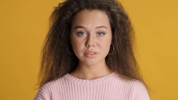 リングイヤリングとピンクのセーターを身に着けているふわふわの髪のブルネットの女の子は 黄色の背景の上にカメラで熱心に見えます 表情コンセプト — ストック動画
