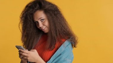 Oldukça pozitif esmer bir kız kameraya bakıyor ve sarı arka planda izole edilmiş akıllı telefondan erkek arkadaşıyla gülüyor.