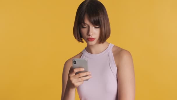漂亮的黑发女孩有着蓬乱的头发 她在社交媒体上被黄色背景隔离的智能手机所吸引 现代技术概念 — 图库视频影像