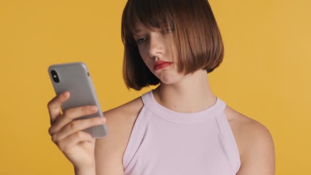 头发蓬乱红唇的黑发女孩在智能手机上浏览社交网络 毫无兴趣 背景是黄色的 悲伤的表情 — 图库视频影像