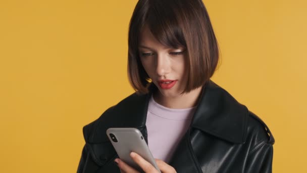 漂亮的黑发女孩用智能手机愉快地同意用黄色背景的相机隔离 — 图库视频影像