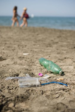 İnsanlar arka planda yürüyen ile kumlu bir plaj üzerinde yeşil, plastik şişe, pamuk temizleme bezi, kap, kutu ve sigara yaptı.