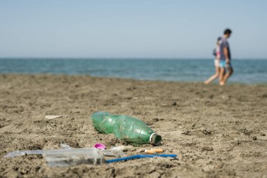 İnsanlar arka planda yürüyen ile kumlu bir plaj üzerinde yeşil, plastik şişe, pamuk temizleme bezi, kap, kutu ve sigara yaptı.