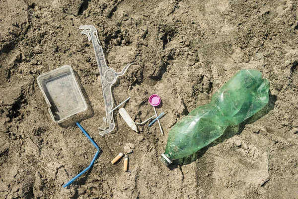 Green, plastic bottle, cotton swabs, cap, box and cigarettes left on a sandy beach.