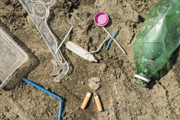 Green, plastic bottle, cotton swabs, cap, box and cigarettes left on a sandy beach.