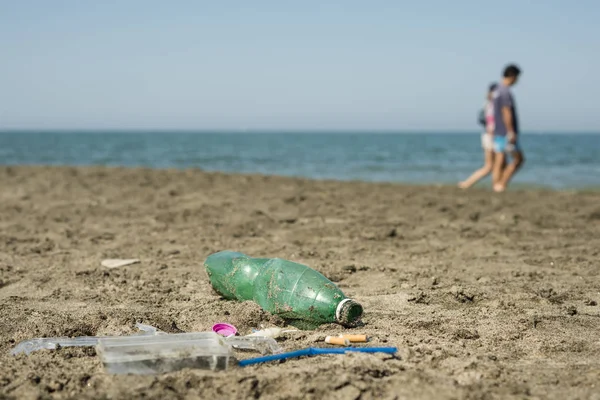 İnsanlar arka planda yürüyen ile kumlu bir plaj üzerinde yeşil, plastik şişe, pamuk temizleme bezi, kap, kutu ve sigara yaptı.