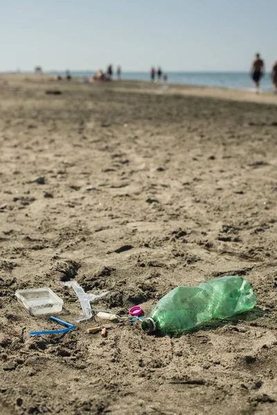 Green, plastic bottle, cotton swabs, cap, box and cigarettes left on a sandy beach with walking people in background.