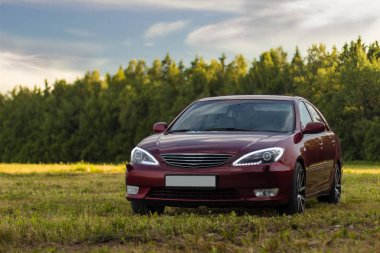 Kiraz kırmızı 4 kapılı aile d-sınıfı sedan Toyota Camry beşinci nesil xv30 2001-2006 led farlar ve 18 inç jantlar büyük jantlar ile yaz bir arka plan üzerinde yeşil bir alan ve orman üzerinde ayakta