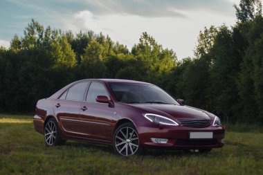 Kiraz kırmızı 4 kapılı aile d-sınıfı sedan Toyota Camry beşinci nesil xv30 2001-2006 led farlar ve 18 inç jantlar büyük jantlar ile yaz bir arka plan üzerinde yeşil bir alan ve orman üzerinde ayakta
