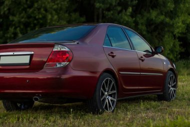 Kiraz kırmızı 4 kapılı aile d-sınıfı sedan Toyota Camry beşinci nesil xv30 2001-2006 led farlar ve 18 inç jantlar büyük jantlar ile yaz bir arka plan üzerinde yeşil bir alan ve orman üzerinde ayakta