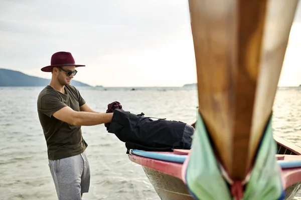 Uomo con stoppie con zaino nero vicino barca di legno — Foto Stock