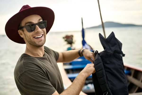 Cheerful man with stubble is holding his backpack near wooden boat — Stock Photo, Image