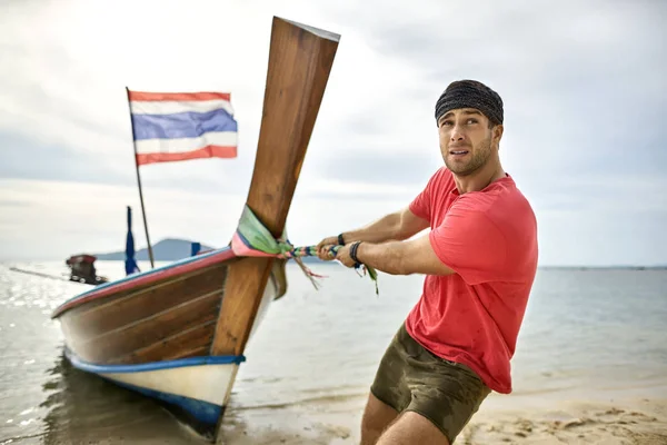 Man with stubble is pulling wooden boat by rope on sand beach — Stock Photo, Image