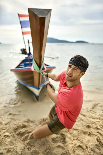 Man with stubble is pulling wooden boat by rope on sand beach