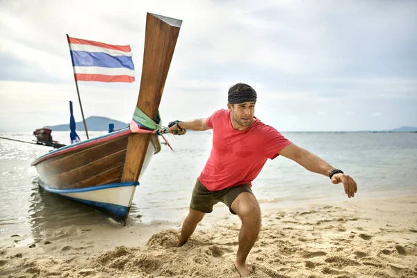 Hombre con rastrojo está tirando de barco de madera por la cuerda en la playa de arena — Foto de Stock