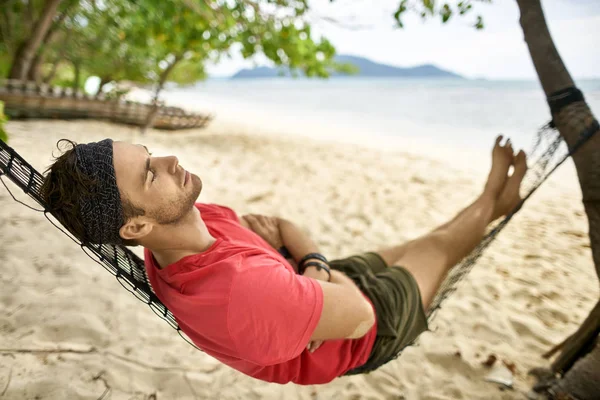 Man with stubble is lying on black hammock on sand beach