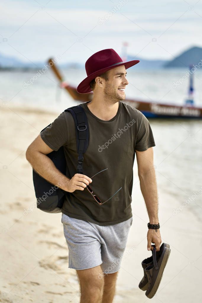 Barefoot man with stubble and backpack is walking on sand beach