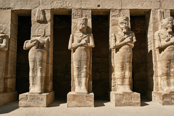 Row of ancient columns with human sculptures on the facade of Karnak Temple in Egypt. Sun shines onto them. Horizontal.