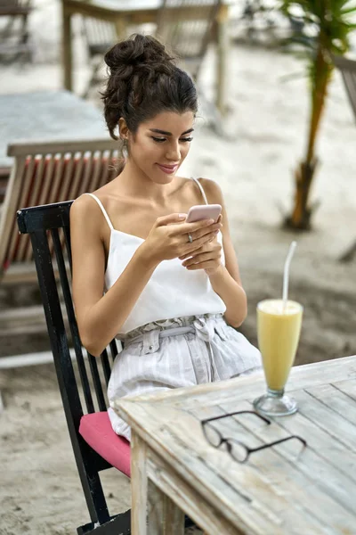 Засмагла жінка використовує свій смартфон у відкритому ресторані на тропічному пляжі — стокове фото