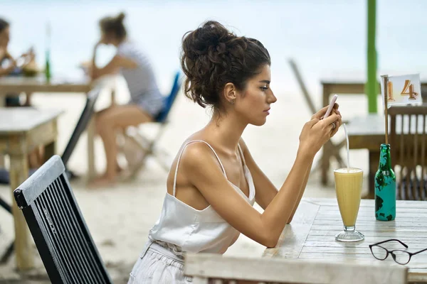 Mujer bronceada usando su teléfono inteligente en el restaurante al aire libre en la playa tropical — Foto de Stock