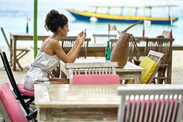 Gebruinde vrouw met behulp van haar smartphone in openlucht restaurant op tropisch strand — Stockfoto
