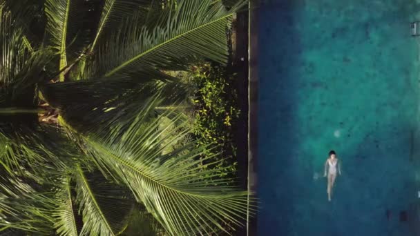 Брюнетка плавает в открытом бассейне возле тропических растений и деревьев — стоковое видео