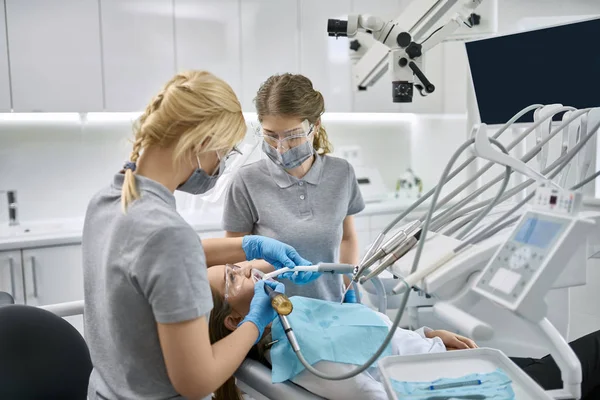 Docteur En Gants Médicaux Noirs Examine Les Dents De La Jeune Fille En  Utilisant Un Microscope Dentaire Et Un Miroir Dans Le Bureau Du Dentiste.  La Femme A Un Bavoir Du Patient