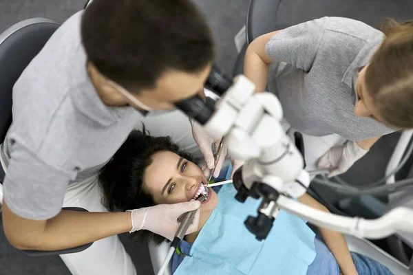 Docteur En Gants Médicaux Noirs Examine Les Dents De La Jeune Fille En  Utilisant Un Microscope Dentaire Et Un Miroir Dans Le Bureau Du Dentiste.  La Femme A Un Bavoir Du Patient