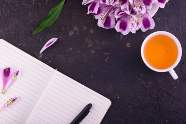 Beyaz bir fincan kokulu çay, güzel çiçekler, siyah bir arka plan, kalem ve gözlük ile bir defter.