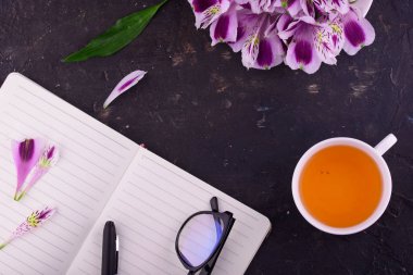 Beyaz bir fincan kokulu çay, güzel çiçekler, siyah bir arka plan, kalem ve gözlük ile bir defter.